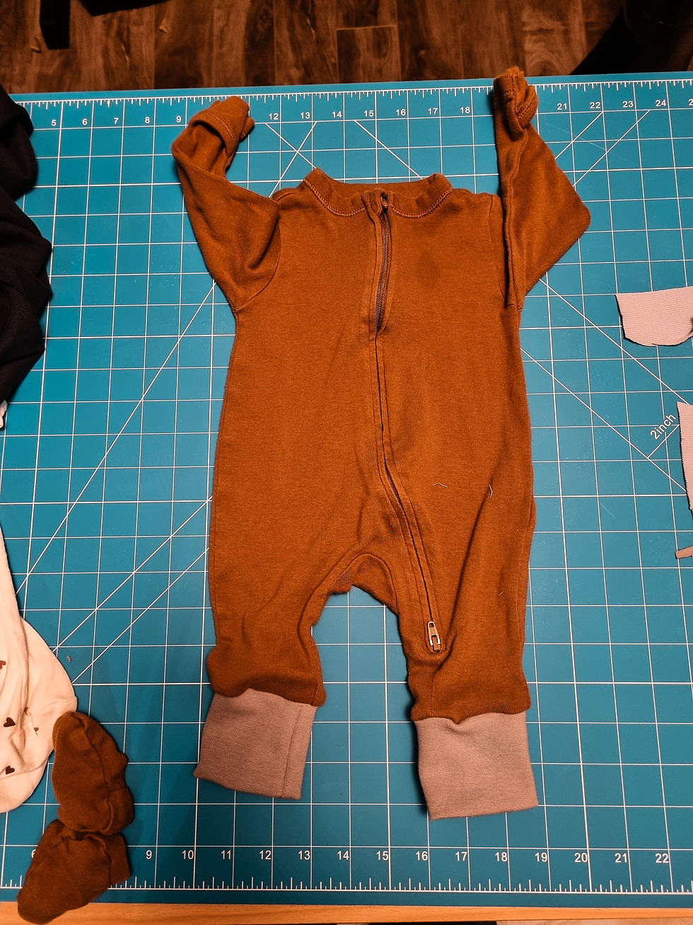 Brown baby onesie with a zipper on a blue cutting mat grid. Beige cuffs, white fabric scraps, and wooden floor visible in the background.