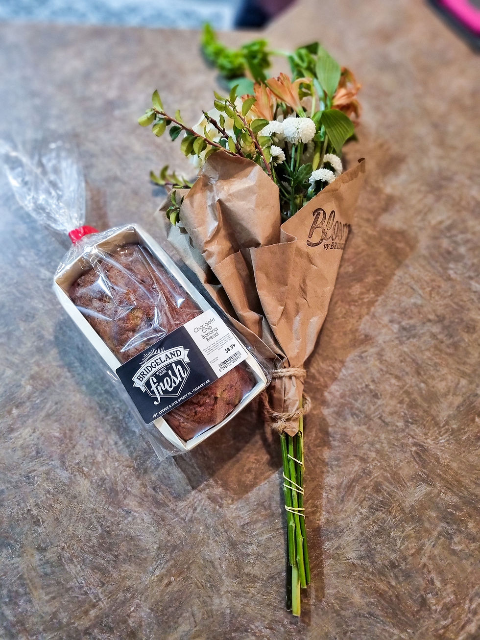 Packaged loaf labeled "Bridgeland Fresh" and a bouquet wrapped in brown paper on a marble table. Bright flowers and greens are visible.
