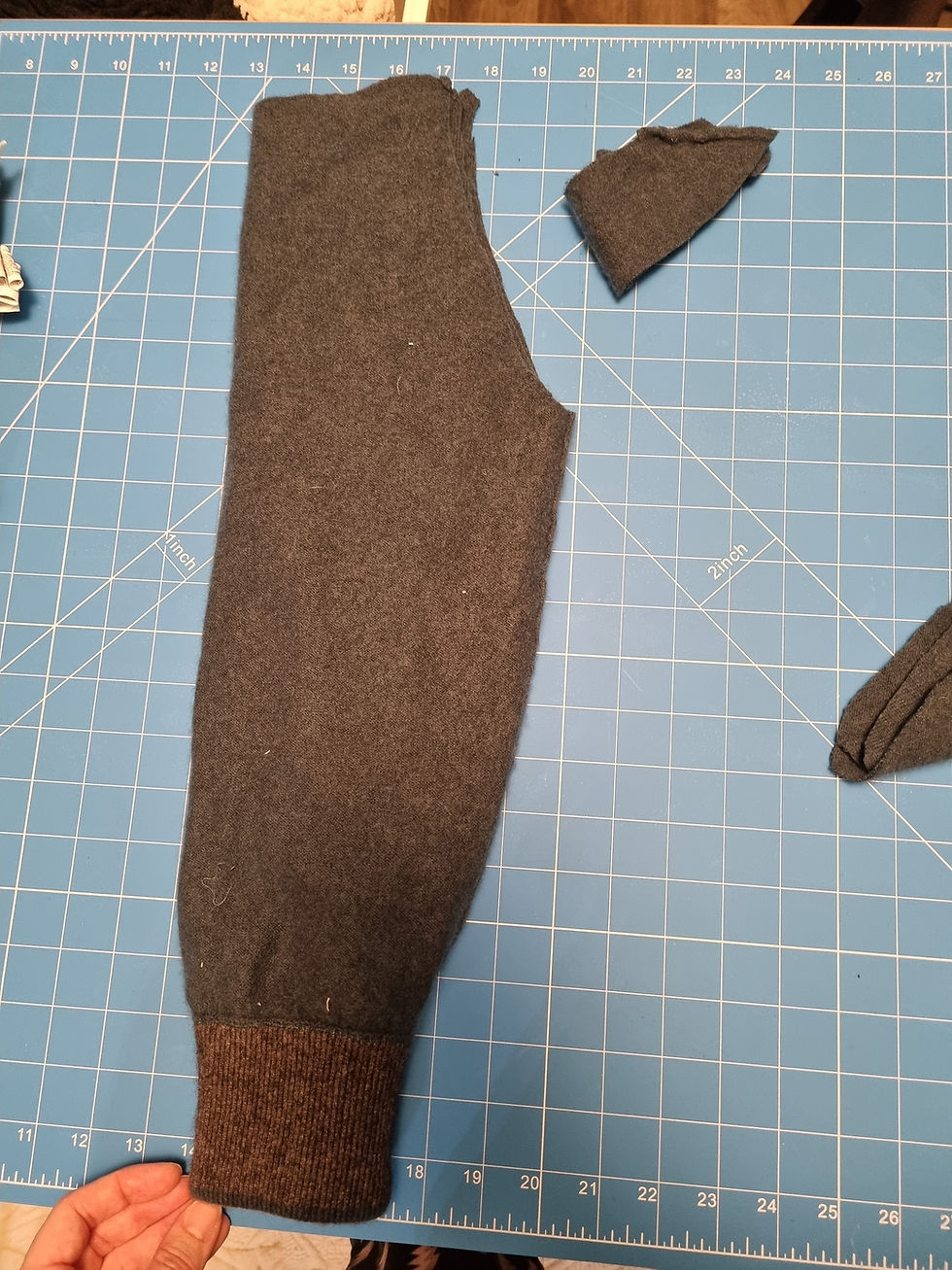 Gray sweater sleeve on a blue cutting mat, with square measurements. A hand holds the cuff, and a fabric piece is cut off beside it.