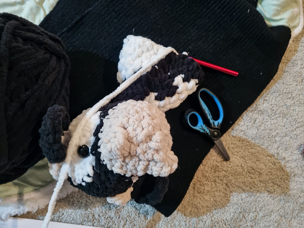 Plush penguin in progress with black, white chunky yarn, red crochet hook, and black scissors on a beige carpet background.