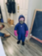 Child in a blue snowsuit and pink mittens stands indoors. Chalkboard and coats in background. Cozy winter scene with playful vibe.