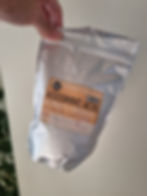 Hand holding silver bag labeled "Ascorbic Acid, Vitamin C Granules." Text includes weight, origin. Background has green plant.