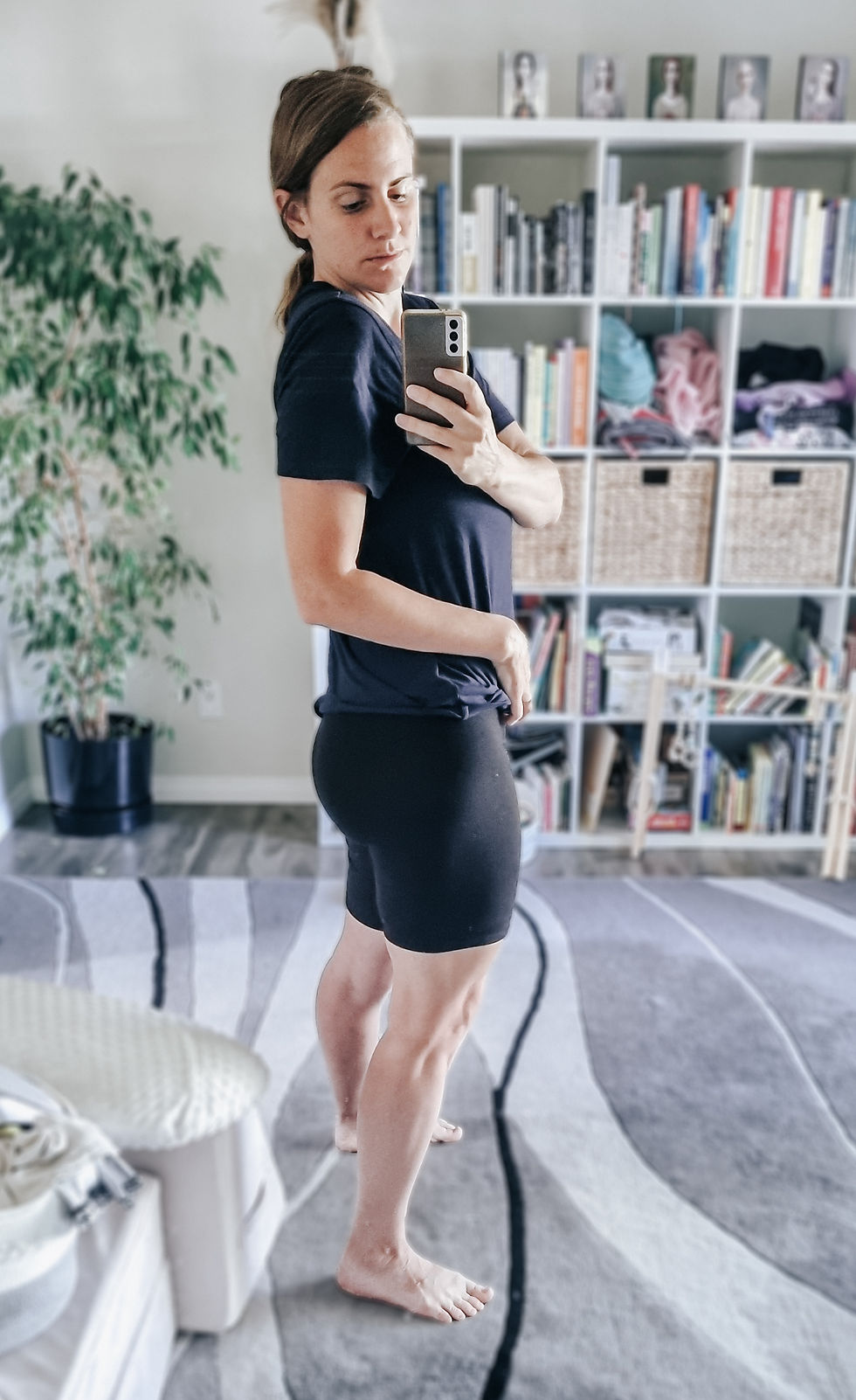 Woman in black athletic clothes takes a mirror selfie in a bright living room with a bookshelf and potted plant.
