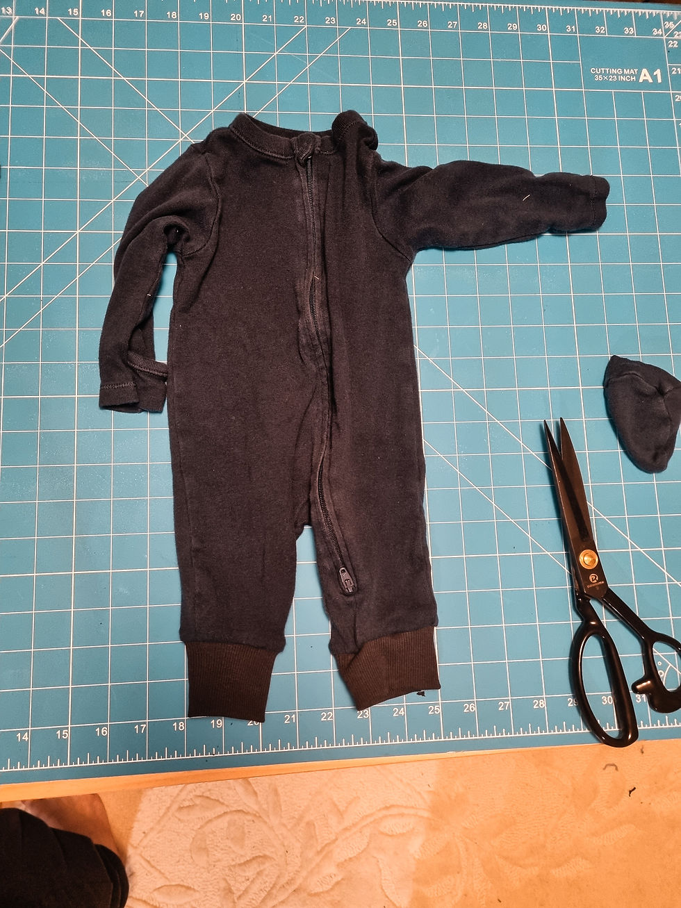 Black baby onesie and scissors on a teal cutting mat with grid lines, showing dimensions. Beige patterned floor visible at the bottom.