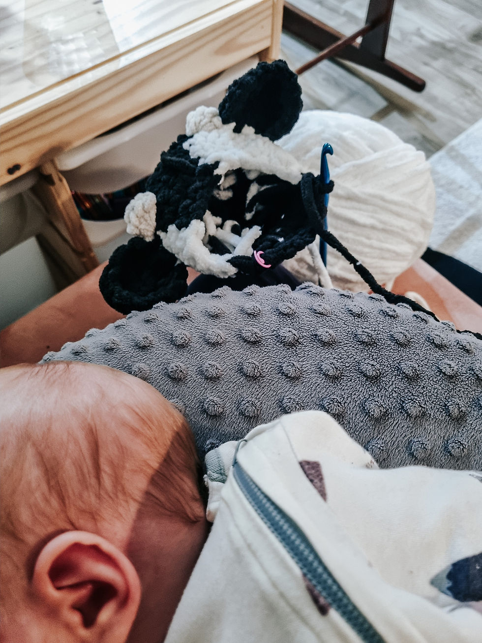 A baby rests on a gray textured pillow next to black and white yarn and a crochet hook, with wooden furniture in the background.