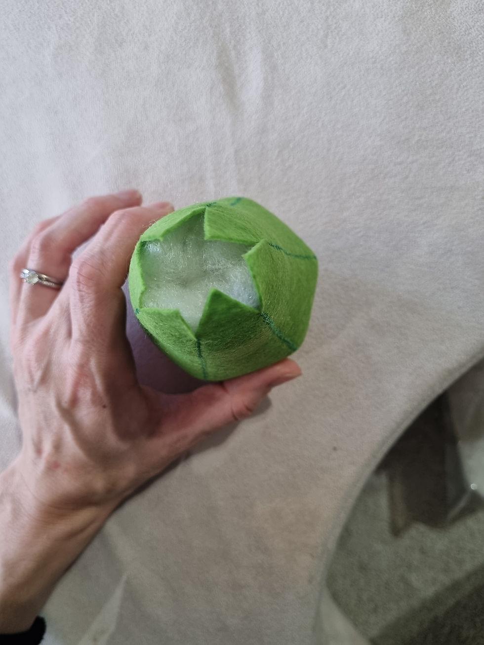 A hand wearing a ring holds a green, partially open felt flower with white fluff inside, against a light fabric background.