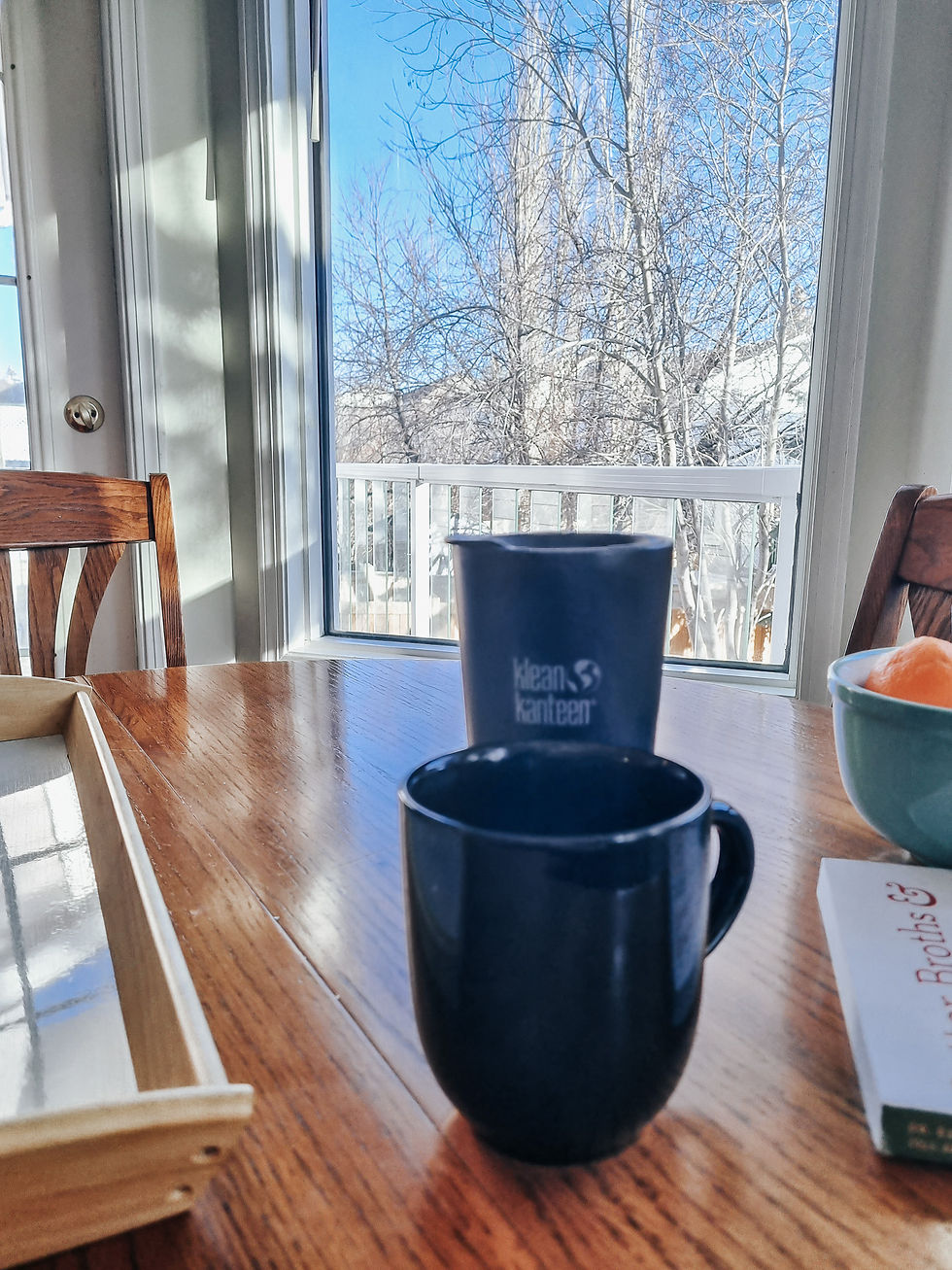 Two black mugs on a wooden table by a window, with a clear blue sky and bare trees outside. A book and fruit bowl nearby. Cozy atmosphere.