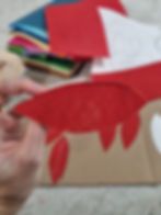 Hand holding red felt leaf, with colorful felt sheets and paper sketches in the background on a gray surface, suggesting a craft project.