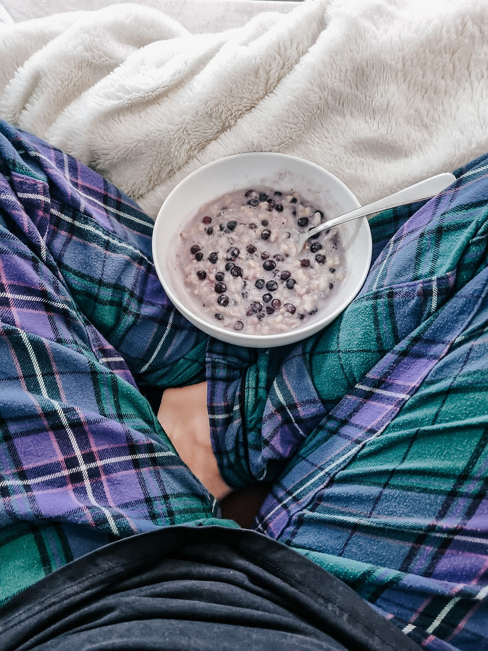 Bowl of oatmeal with blueberries and a spoon on plaid pajamas. Cozy, relaxed setting with a soft cream blanket in the background.