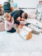 A man kneels holding a baby, while a young girl gestures nearby. They're in a cozy, cluttered living room with toys and a fluffy chair.