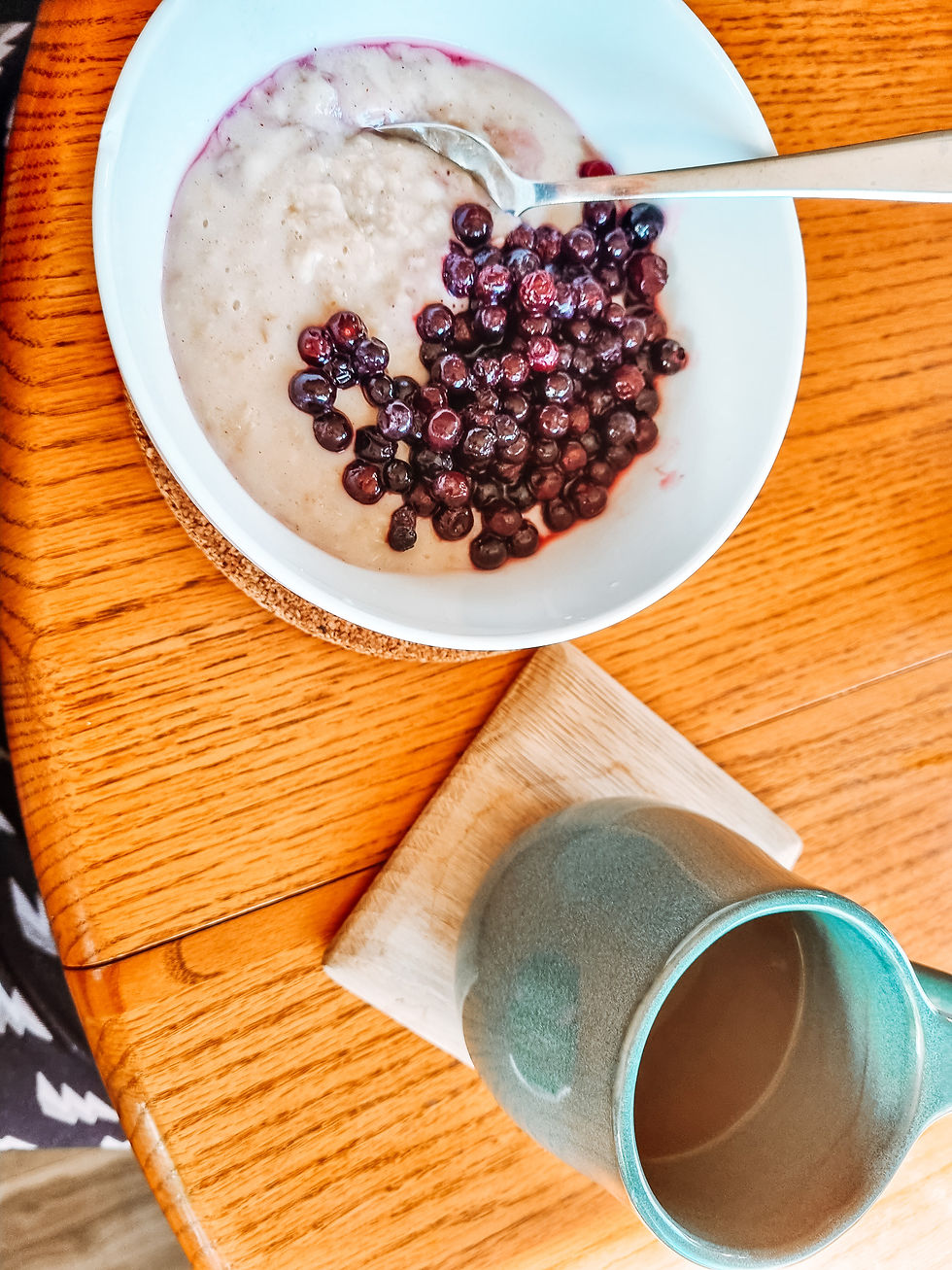 A white bowl of oatmeal with blueberries and a spoon on a wooden table, next to a teal mug on a coaster. Cozy breakfast setting.