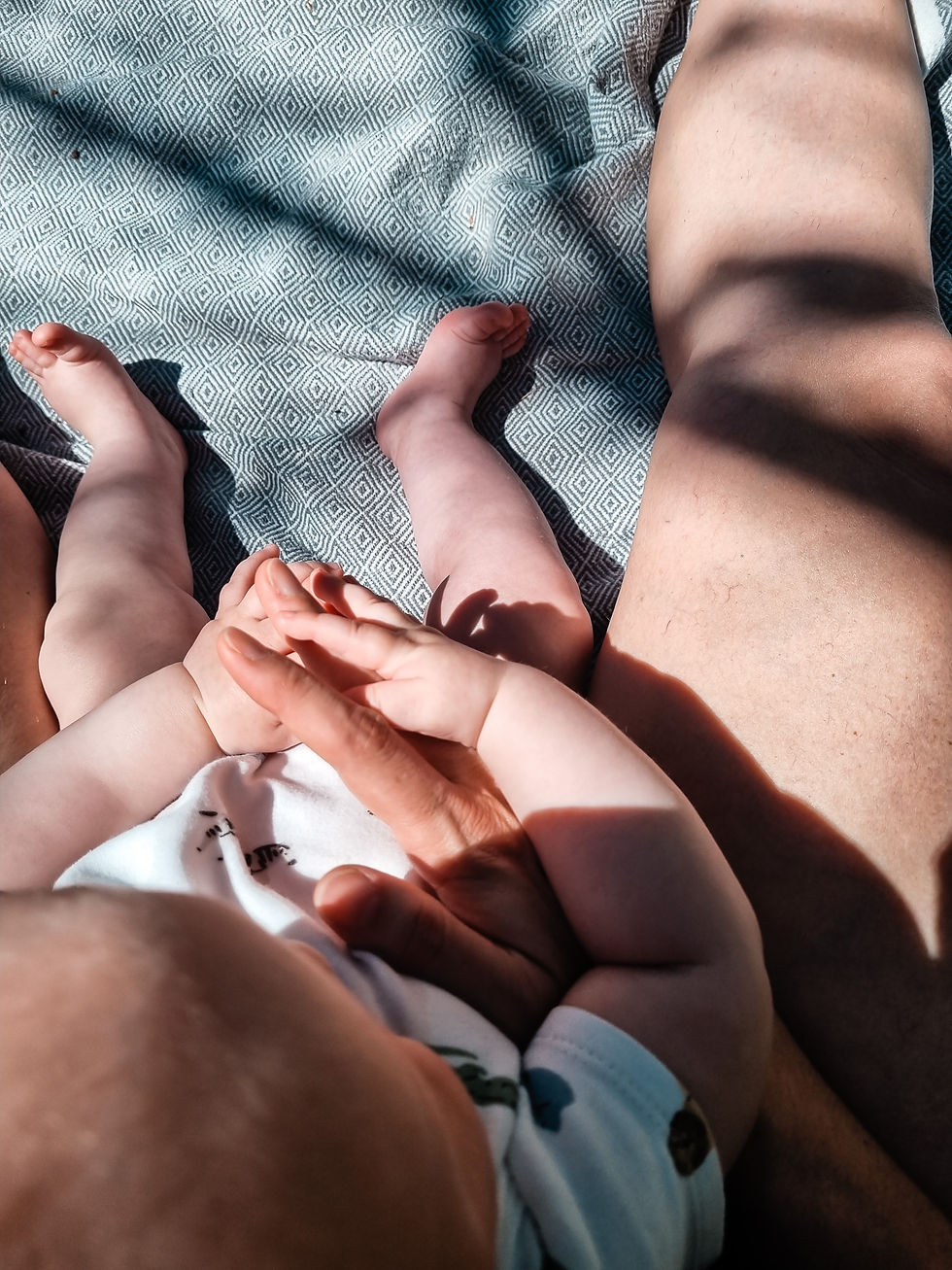 Baby lies between two adults on a patterned blanket, holding hands in warm sunlight.
