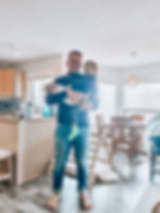 Man holding two children in a bright kitchen, wearing casual clothes. One child wears green socks. Wooden furniture and soft lighting in background.
