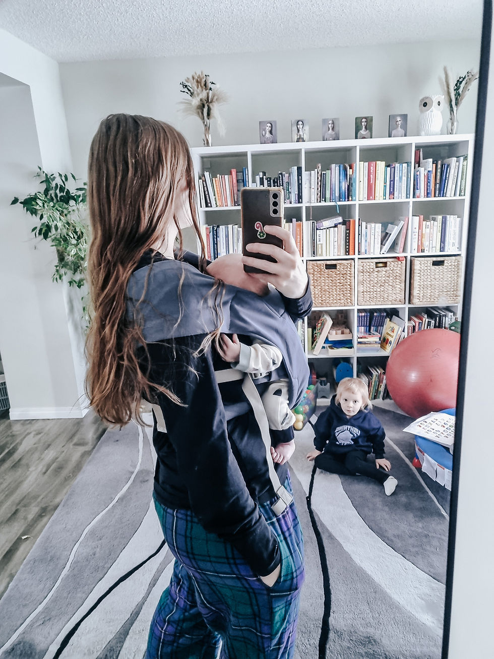 Woman with baby in a carrier takes a mirror selfie. Child sits on a patterned rug in a cozy room with bookshelves and a red exercise ball.