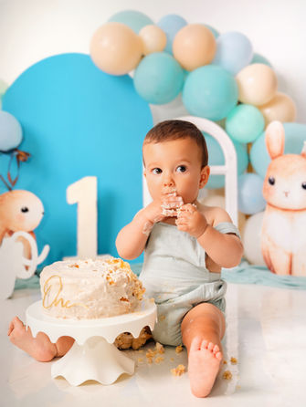 Niño sonriendo y jugando en el suelo, lleva un peto beige. Fondo en tonos azules con globos, dibujos de conejos y decoración "un año". El niño tiene un año, sonrie en la foto . en primer plano se ve la tarta de cumpleaños. Sesión de smash cake en estudio.