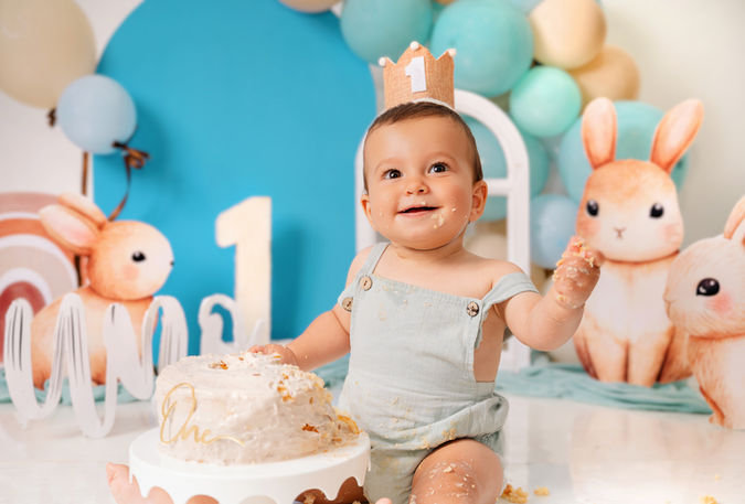 Bebé sonriente con pastel, número 1, rodeado de globos y conejos.
