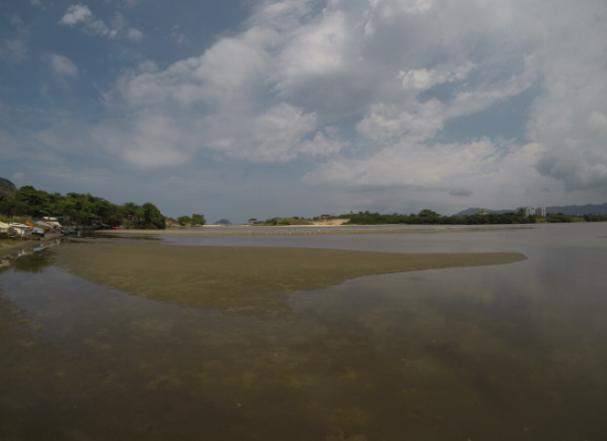 A fotografia mostra uma Laguna sendo vista por sua margem. As margens da lagoas são constituídas por vegetação esverdeada e pode-se observar interferência antrópica. O céu é azul com nuvens.