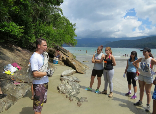 Foto de professor e alunos em curso de Biologia Marinha do projeto Bióicos antes de iniciar uma trilha em área de Mata Atlântica com mar à direita e montanhas ao fundo. Os alunos estão voltados para o professor, que passa instruções.