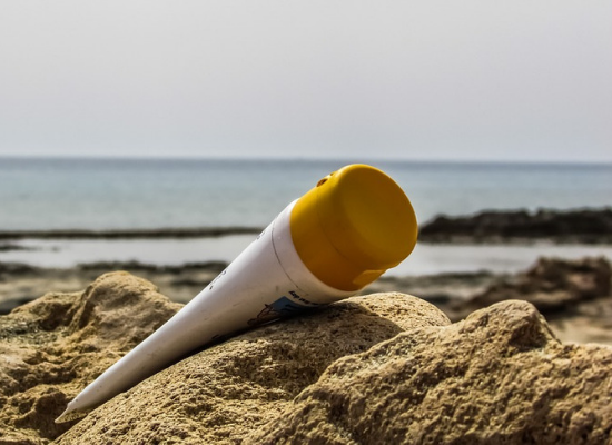 Protetor solar jogado na areia. Ao fundo temos uma imagem desfocada do mar e algumas rochas.