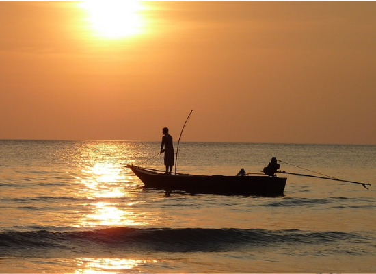 Fotografia da silhueta de um pescador segurando vara de pescar em pé em uma canoa motorizada no mar. Ao fundo, é possível ver o brilho do sol no céu avermelhado.
