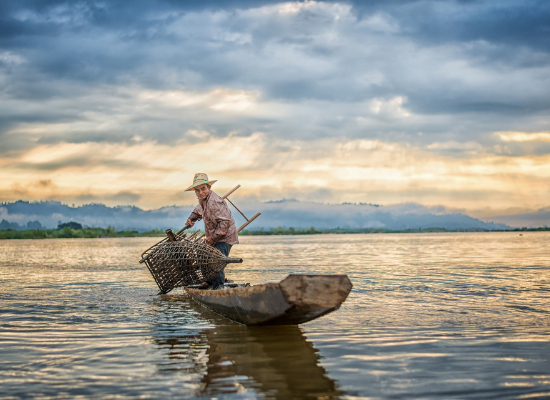 A fotografia mostra em primeiro plano, um homem em cima de um barco simples, com uma armadilha na mão, utilizada para a pesca. Ao fundo estão montanhas e o céu está coberto de nuvens.