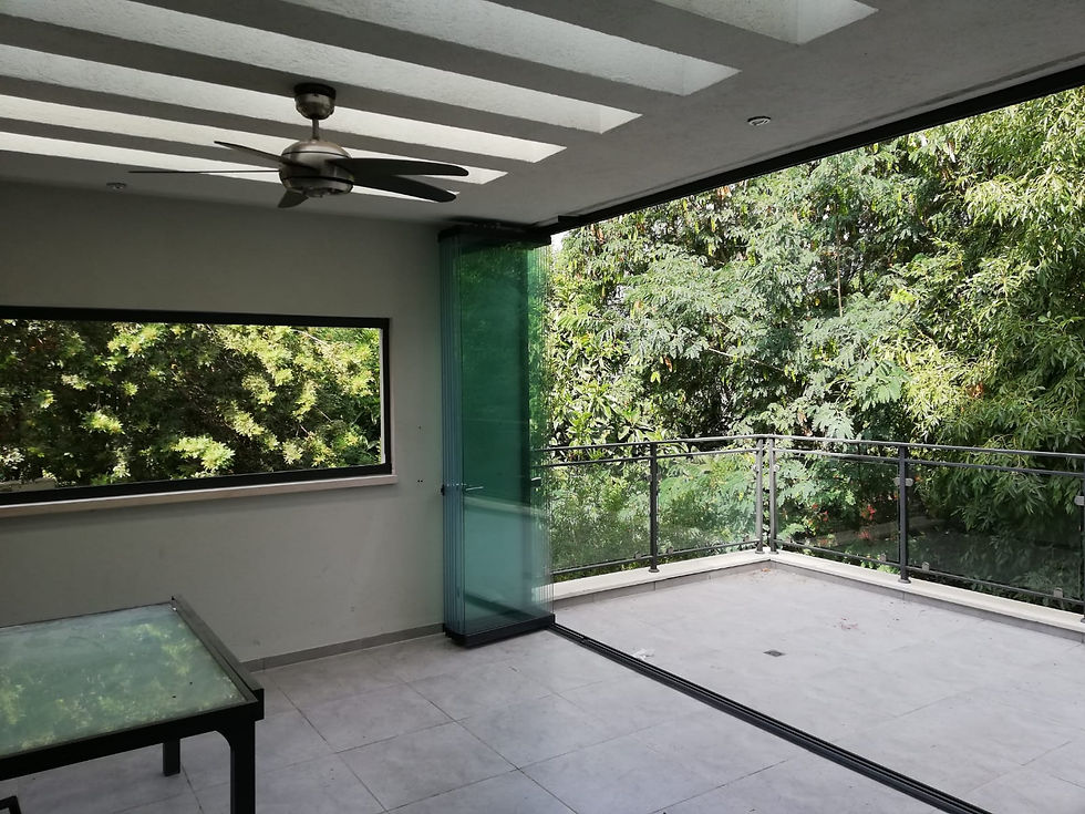 Open balcony with privlux tiara folding glass walls and a ceiling fan, overlooking lush green trees. A glass-topped table stands on a tiled floor. Tranquil setting.