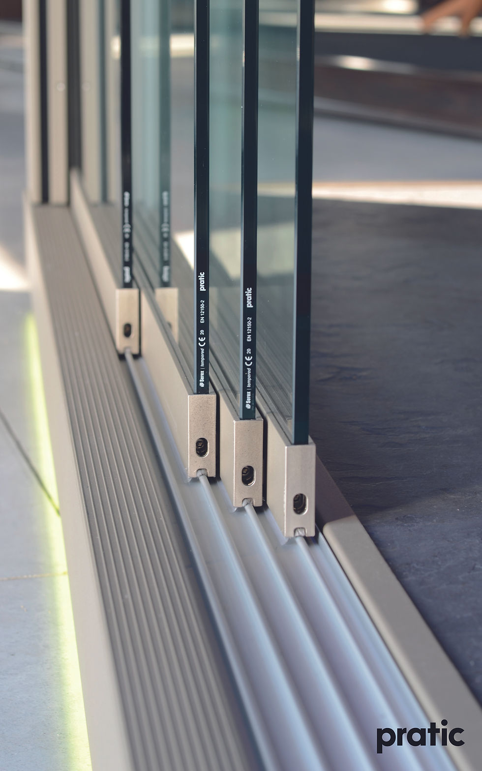 Sliding glass doors with metal hinges on a grooved track. Text "pratic" visible. Modern, sleek design with a light-filled setting.