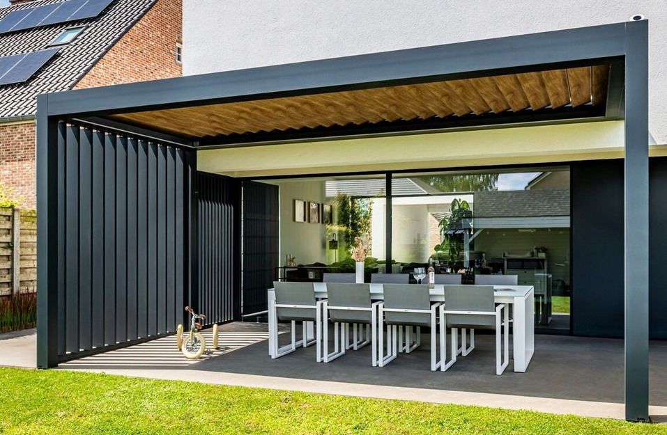 Outdoor patio with modern black pergola, and wooden roof, ambiente vertical louver walls, white dining table, and eight gray chairs. A tricycle is in the corner. Glass doors reflect greenery.