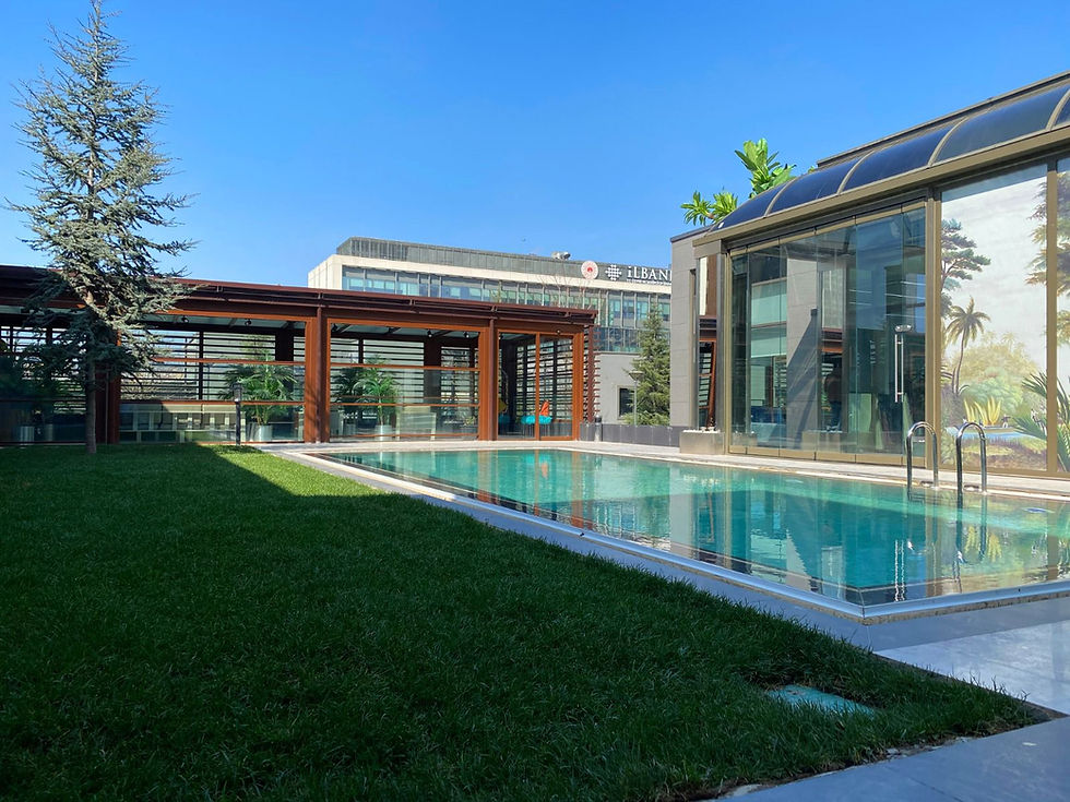 Modern outdoor pool beside glass buildings and green lawn under a clear blue sky, with partial sign text reading ILBAN.