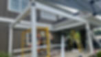 Privlux Workers install a white framed pergola on a brown house. Yellow scaffolding and ladders are around the area. The mood is industrious.