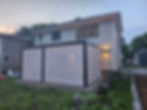 Modern two-story house with a lit pergola enclosed by sheer shades. Toys and chairs outside, surrounded by grass and trees at dusk.