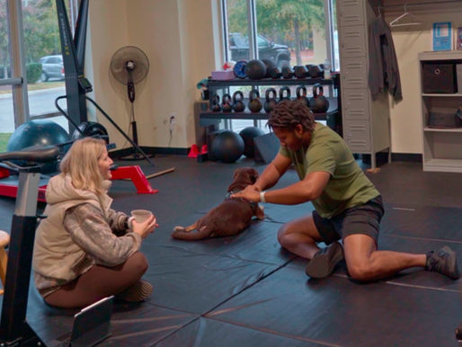 a woman sitting on an exercise mat with a coffee mug, talking to and instructing a man who is performing a hip exercise, and a dog who is laying on the exercise mat looking out the windows of the gym