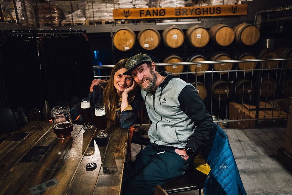 Lächelndes Paar in der Brauerei vor Fässern und dem Schild FANØ BRYGHUS, mit dunklen Biergläsern am Holztisch.