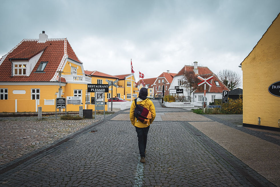 Person mit gelber Jacke und Rucksack geht über gepflasterte Straße in dänischem Ort; gelbe Häuser, Flaggen, Restaurant Plesner.