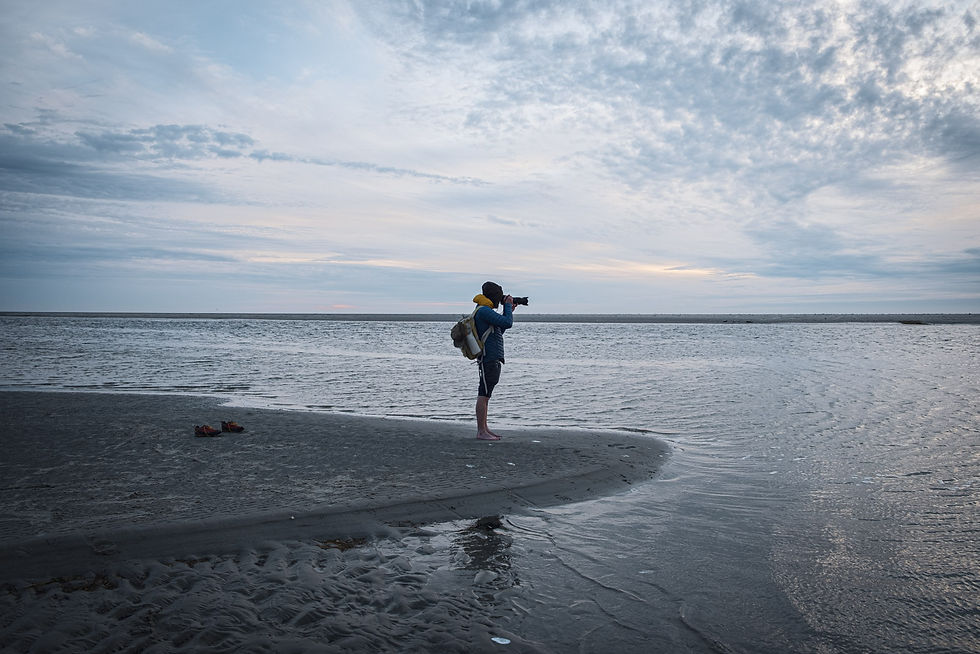 Fotograf mit Rucksack steht barfuß am ruhigen Strand und fotografiert aufs Meer unter wolkigem Morgenhimmel.