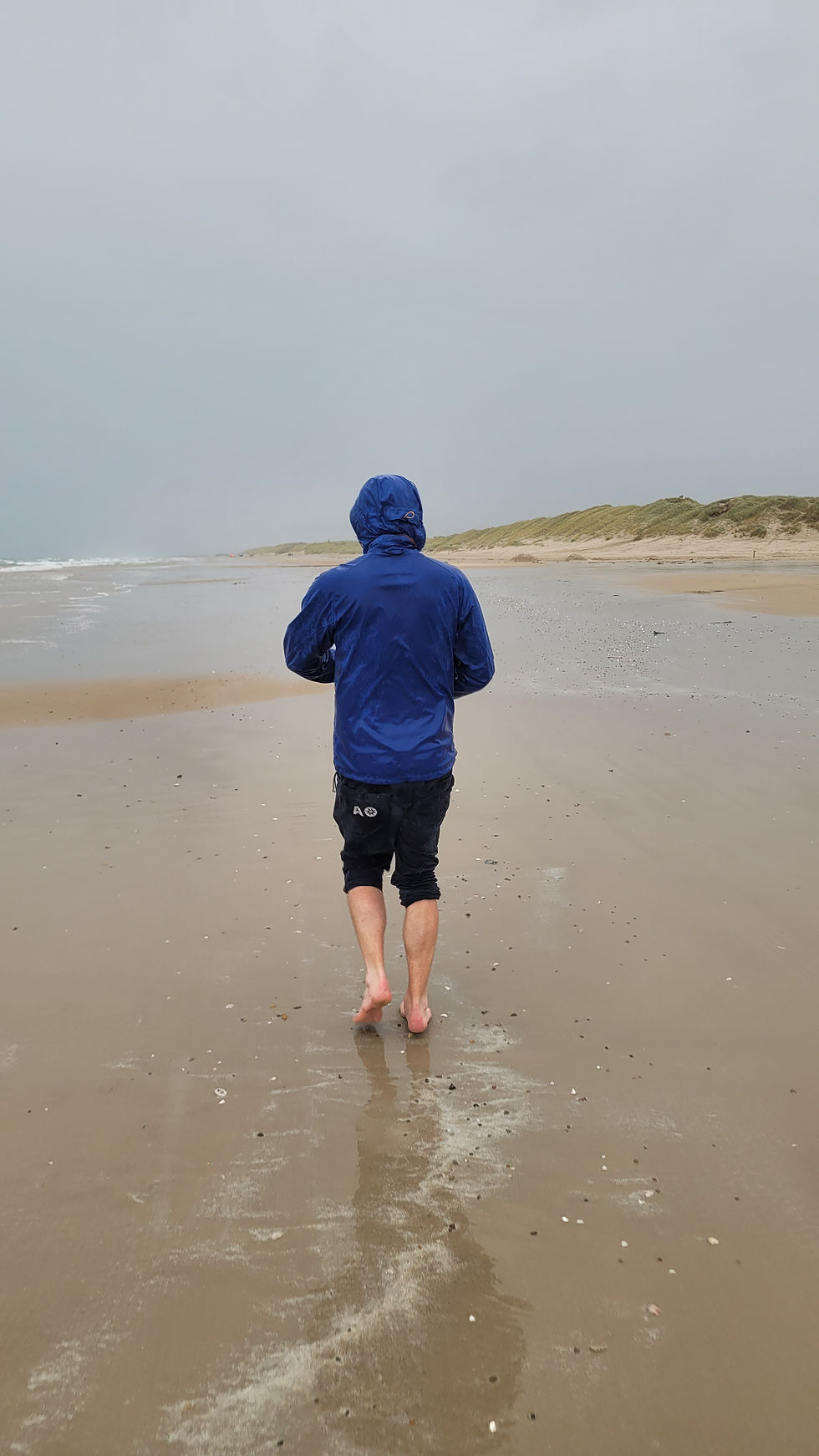 Person in blauer Regenjacke läuft barfuß am leeren Strand unter grauem Himmel.