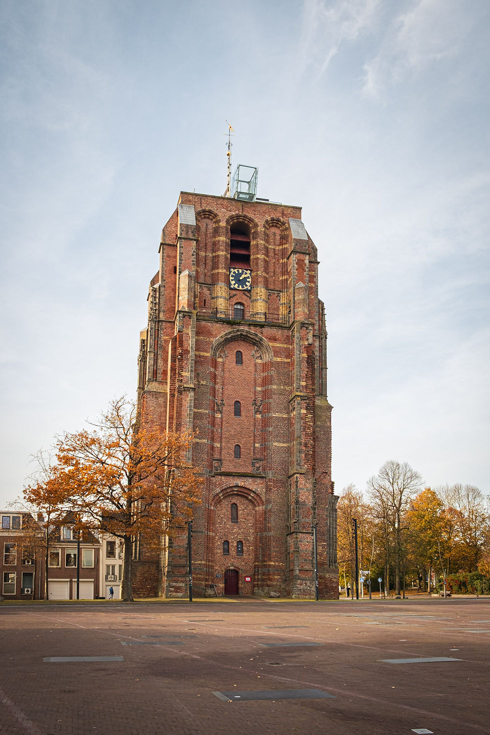 Hoher Backsteinturm mit Uhr auf leerem Platz, umgeben von Herbstbäumen unter hellem Himmel.