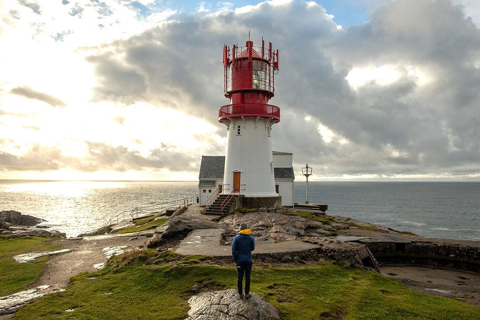 Person vor rot-weißem Leuchtturm an felsiger Küste unter wolkigem Himmel, Blick aufs Meer.