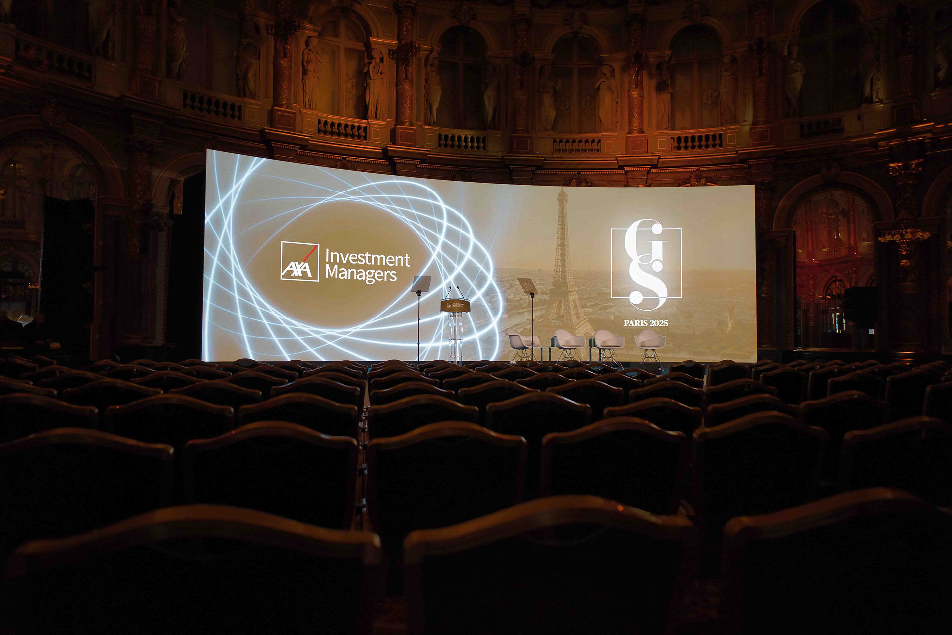 Empty auditorium with ornate architecture, featuring a large screen displaying AXA Investment Managers, G l9 Paris 2025, and the Eiffel Tower.