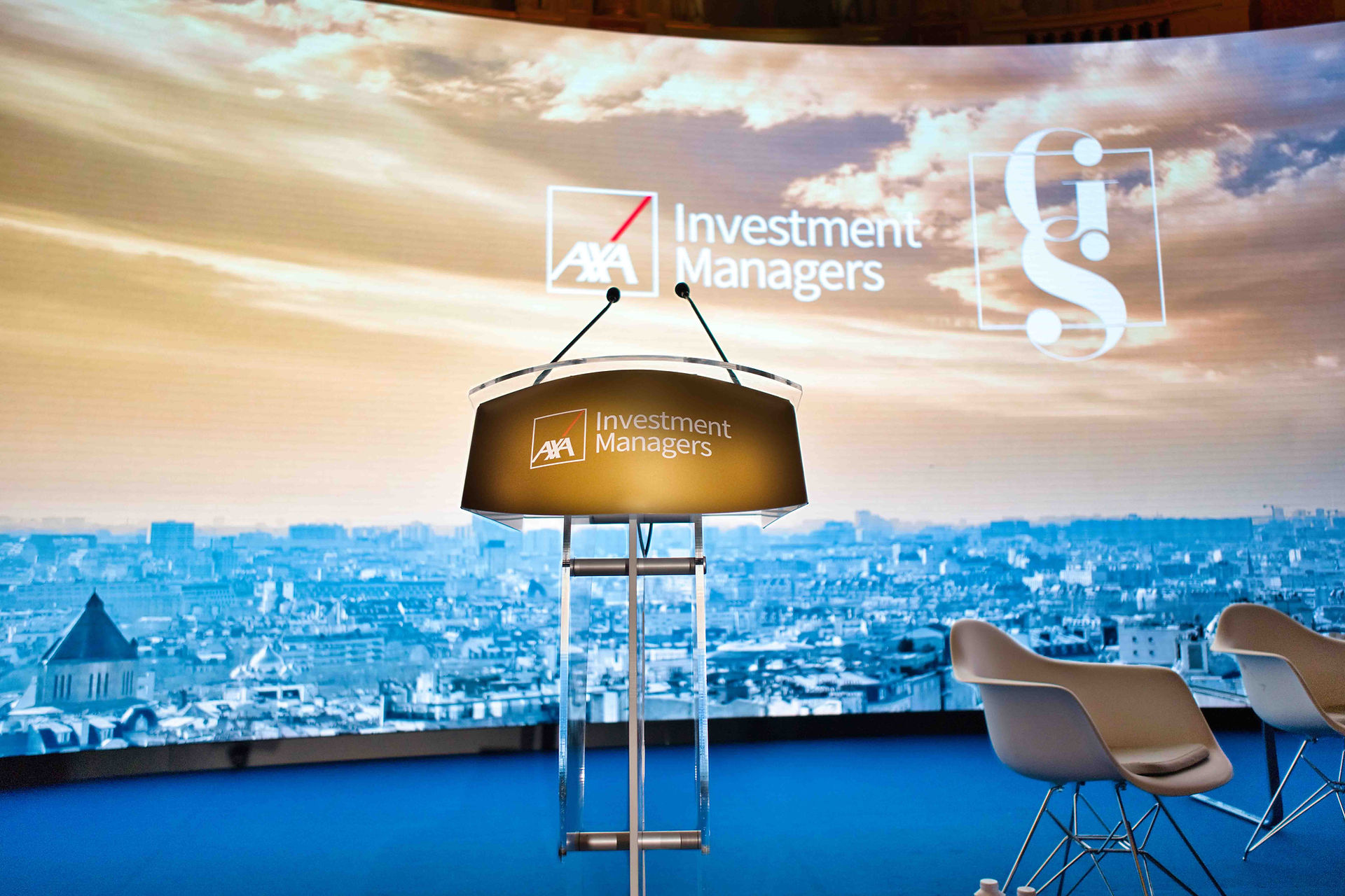 Podium with two microphones on a stage, backdrop showing cityscape and AXA Investment Managers logo. Blue and golden tones dominate.