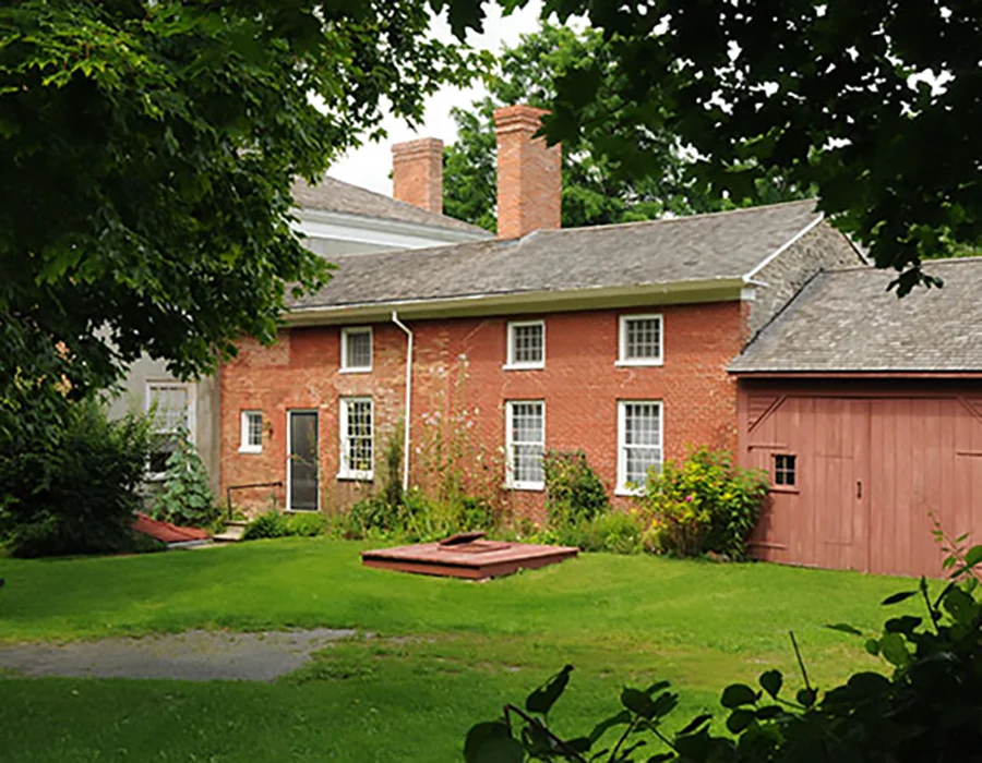 Historic brick house with white windows and chimneys, surrounded by lush greenery and trees, set against a cloudy sky. Quiet and serene mood.