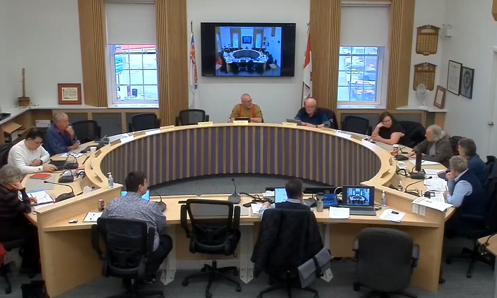 People in a conference room sit around a curved table, engaged in discussion. A screen on the wall displays a similar meeting view.