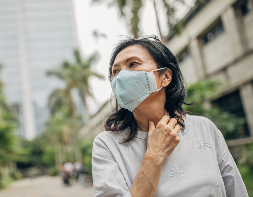 A woman wearing a blue mask looks thoughtful outdoors, surrounded by buildings and trees. She touches her neck, creating a calm mood.