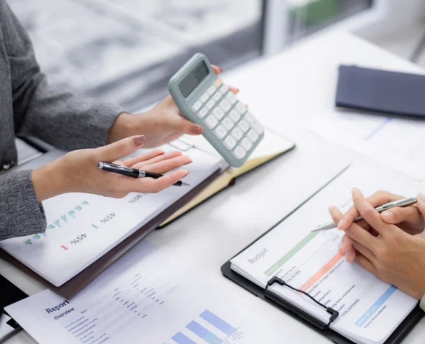 Two people discuss finances at a desk, one holding a calculator, the other a pen. Papers with graphs and "Budget" text are visible.