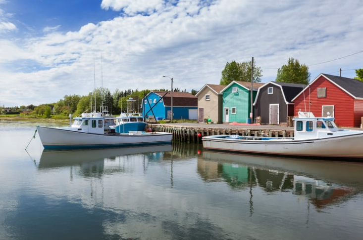 Boats docked at a colorful harbor with red, green, and blue buildings. Calm water reflects the vibrant scene under a partly cloudy sky.