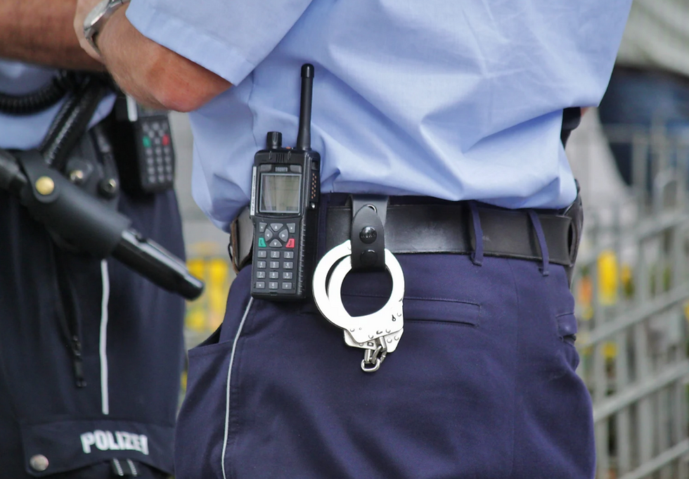 Police officer in blue uniform with radio and handcuffs on belt. "Polizei" text visible. Setting is outdoors, with blurred background.