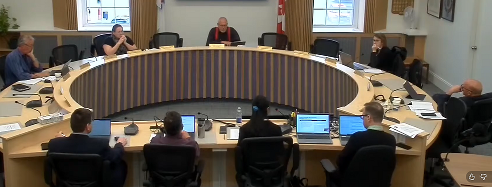 Eight people sit around a curved conference table with laptops and papers. A man in the center speaks. Canadian flag in the background.