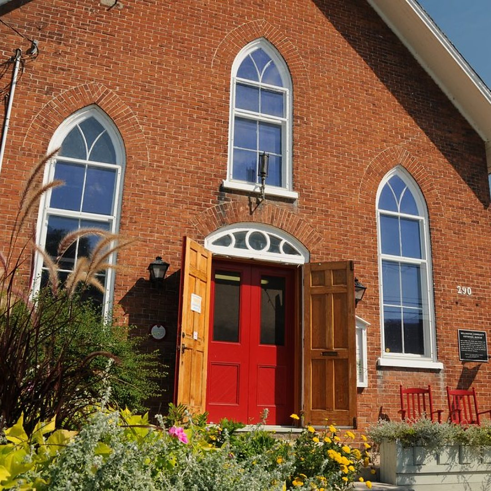 Brick church facade with arched windows, open red double doors, and flower beds in bright sunlight