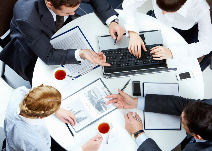 Overhead view of four coworkers around a round table, discussing a laptop and charts with coffee cups in a bright office.