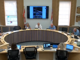 Council meeting in a bright room with people seated around a circular table, using laptops. A presentation is displayed on a screen.